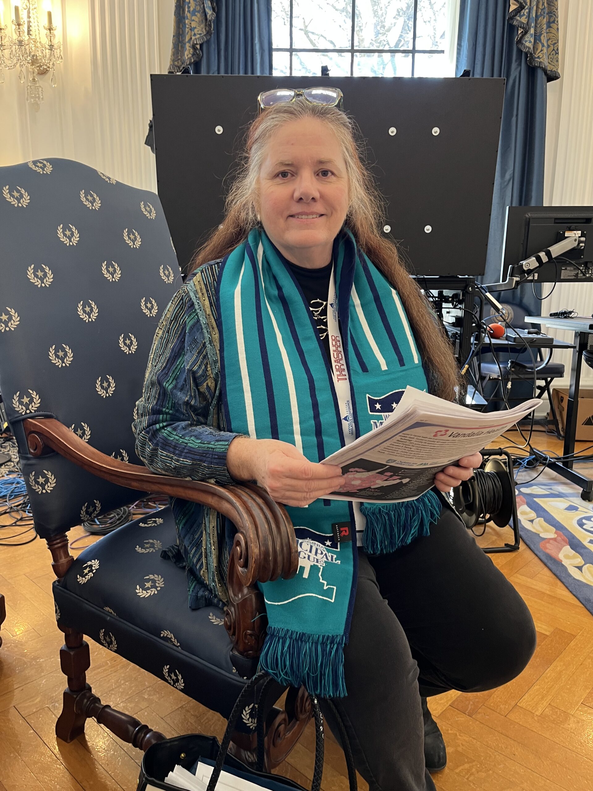 A woman with long hair sits in an ornately carved chair, wearing a teal-striped scarf and reading a newspaper. A tech setup is visible behind her.
