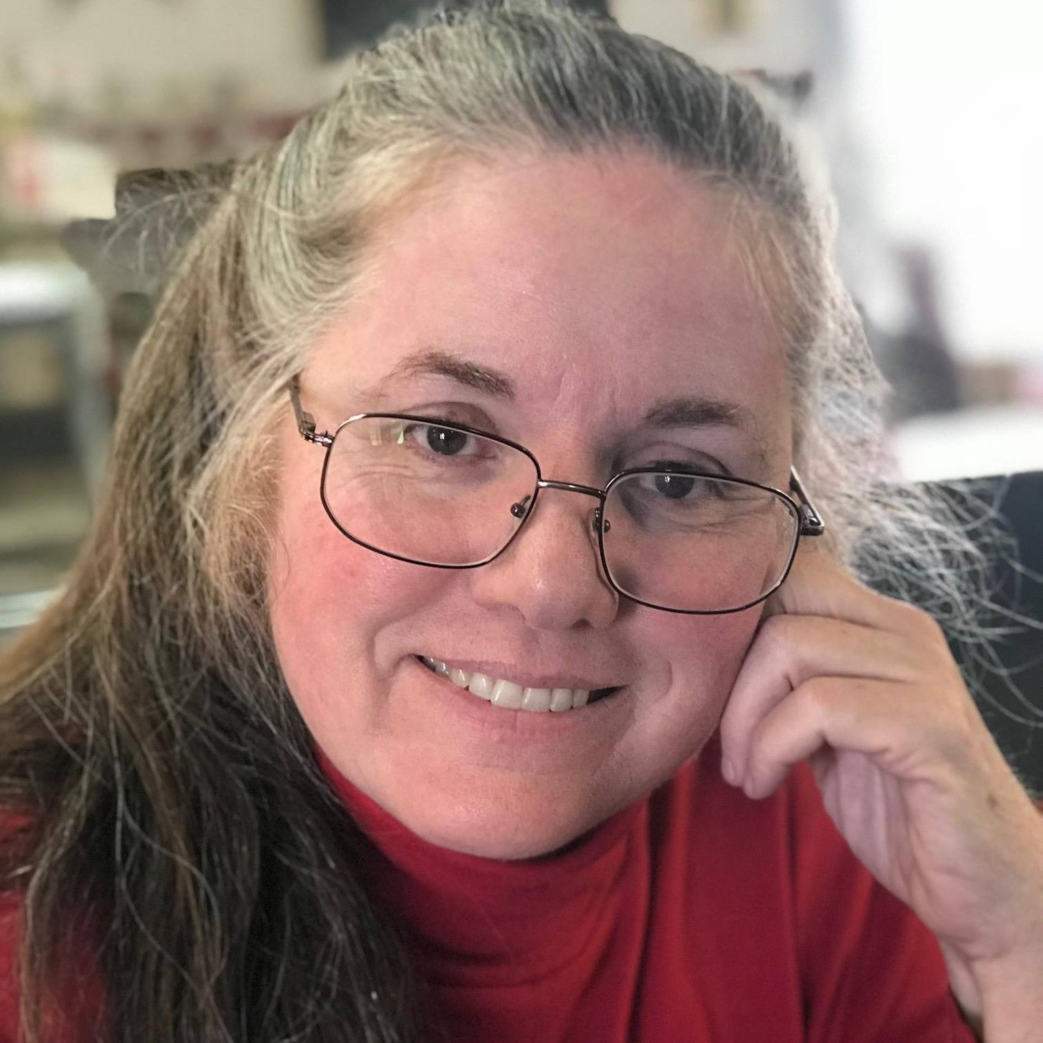 Smiling woman with long gray hair and glasses rests her chin on her hand, wearing a red turtleneck, conveying warmth and approachability.