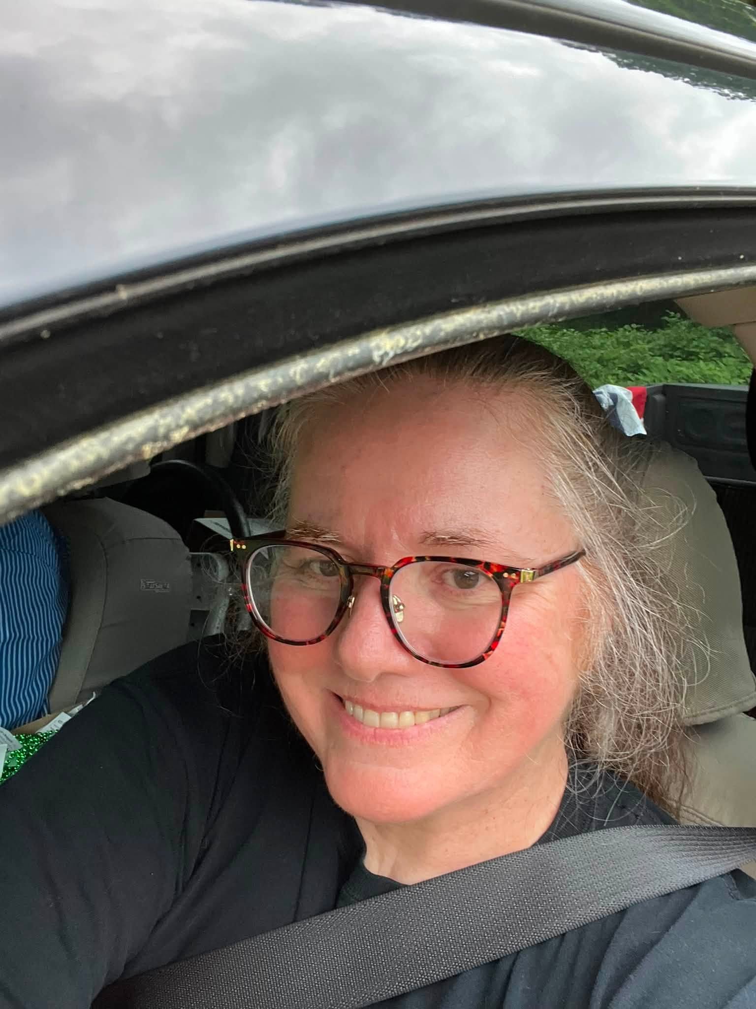 Smiling woman with glasses leans in from a car window, wearing a black shirt. The scene suggests a casual, friendly moment during a drive.