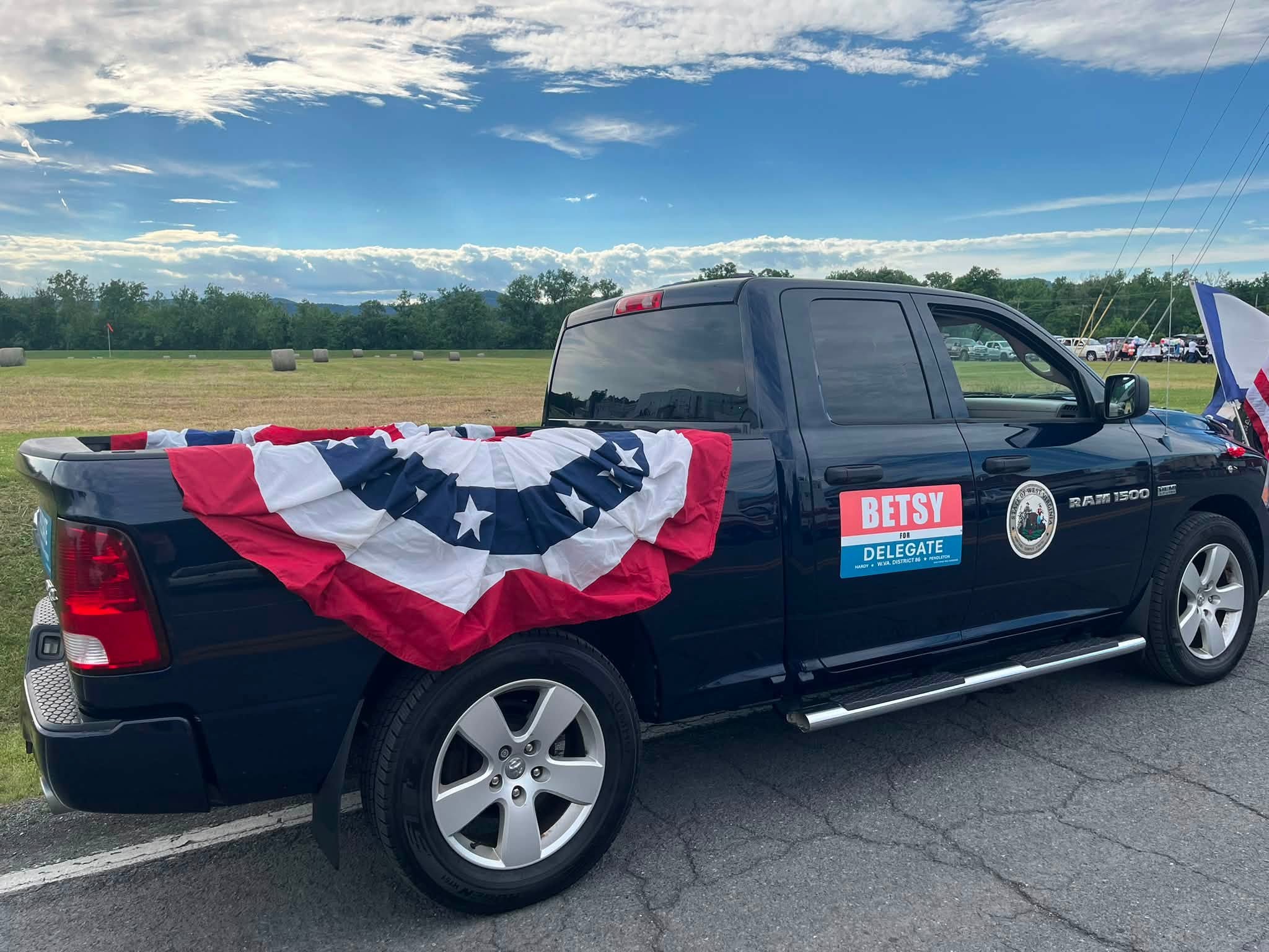 Black pickup truck decorated with a patriotic red, white, and blue bunting and a campaign sign for "Betsy for Delegate." Grass field and sky visible in the background.