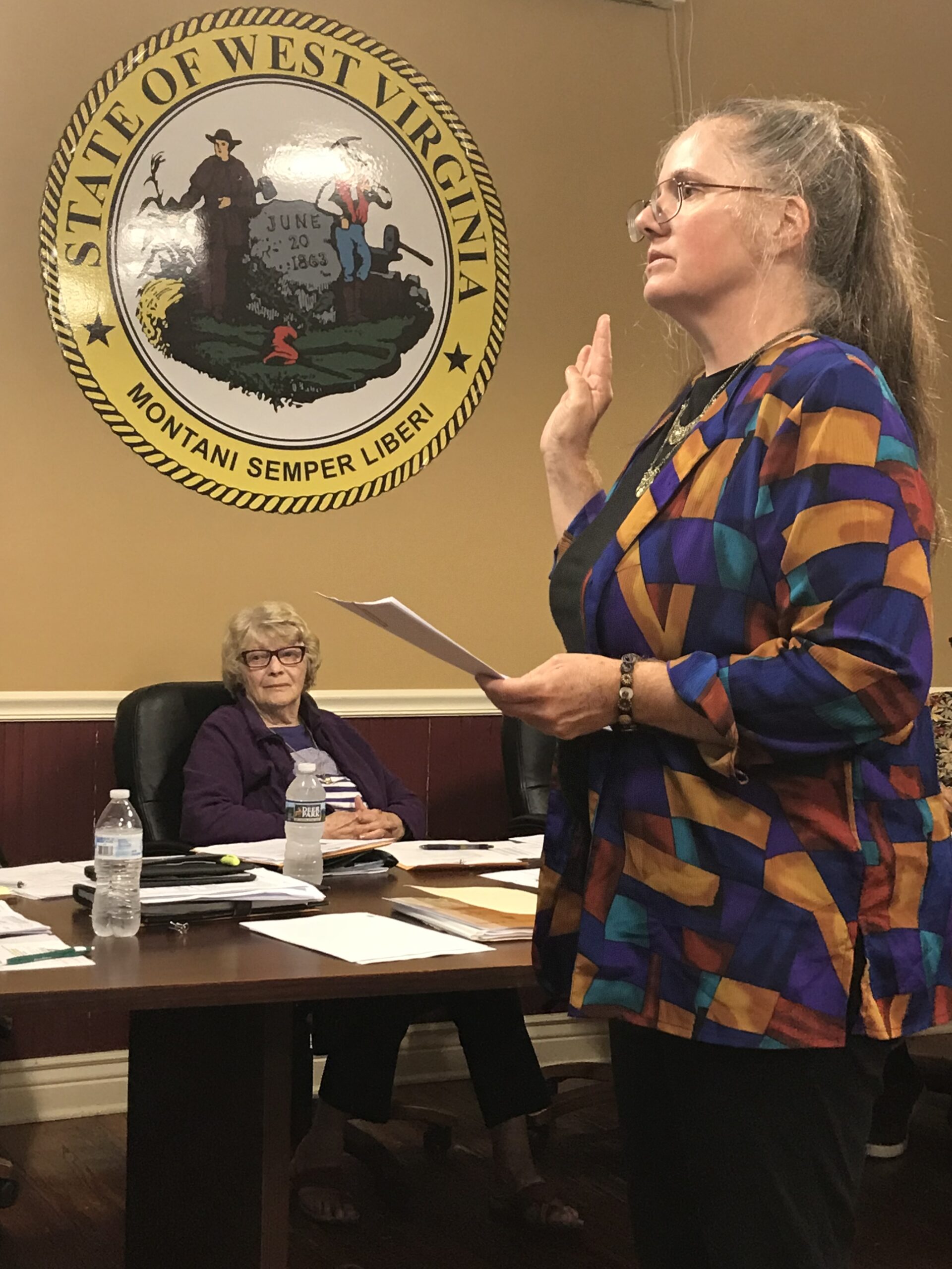 A woman in a colorful jacket is raising her right hand in a formal setting, likely taking an oath, with a state seal of West Virginia displayed behind her.