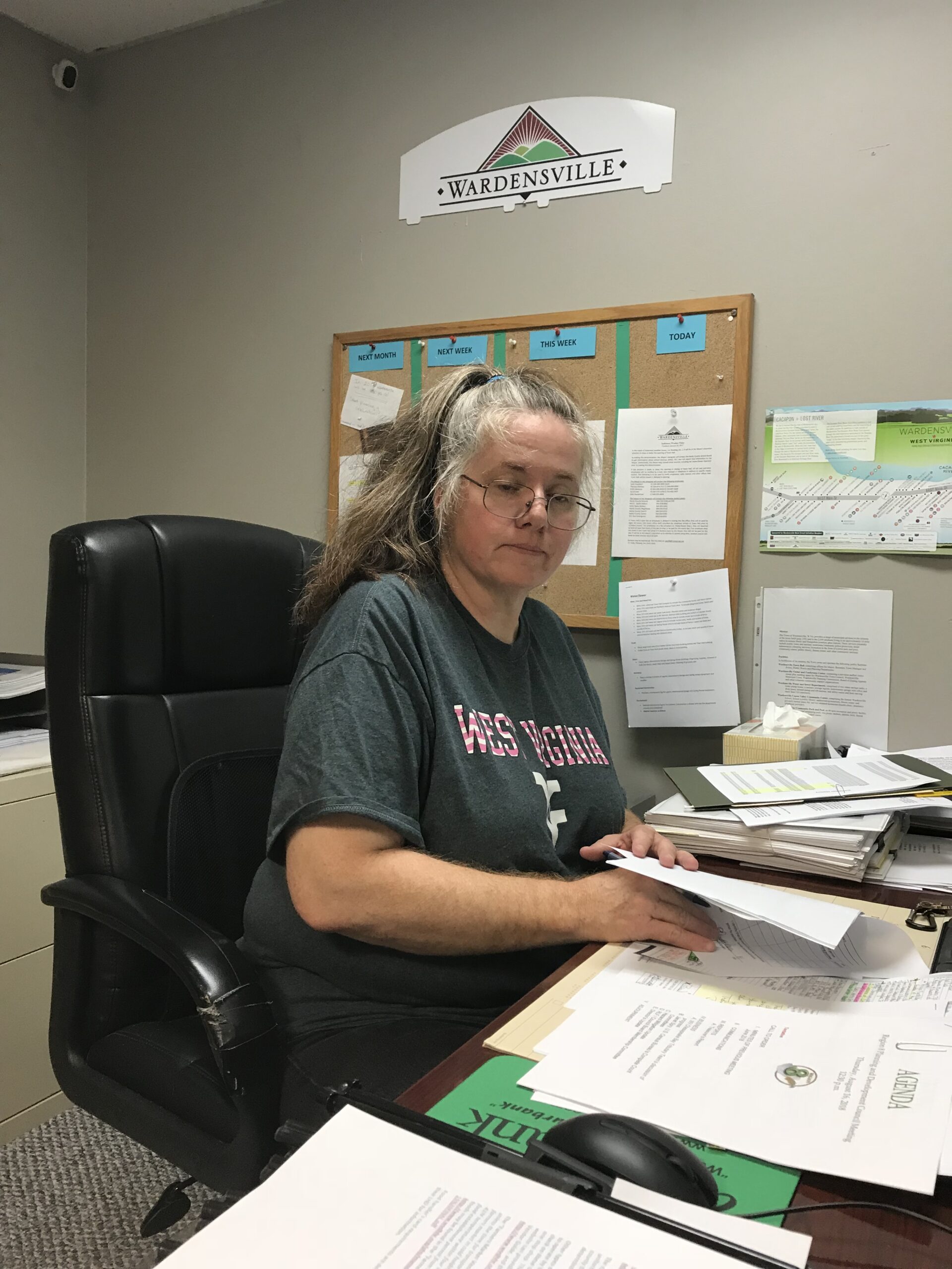 Woman in a "West Virginia" shirt seated at a cluttered desk in an office, reviewing documents. A sign for "Wardensville" is visible above her.