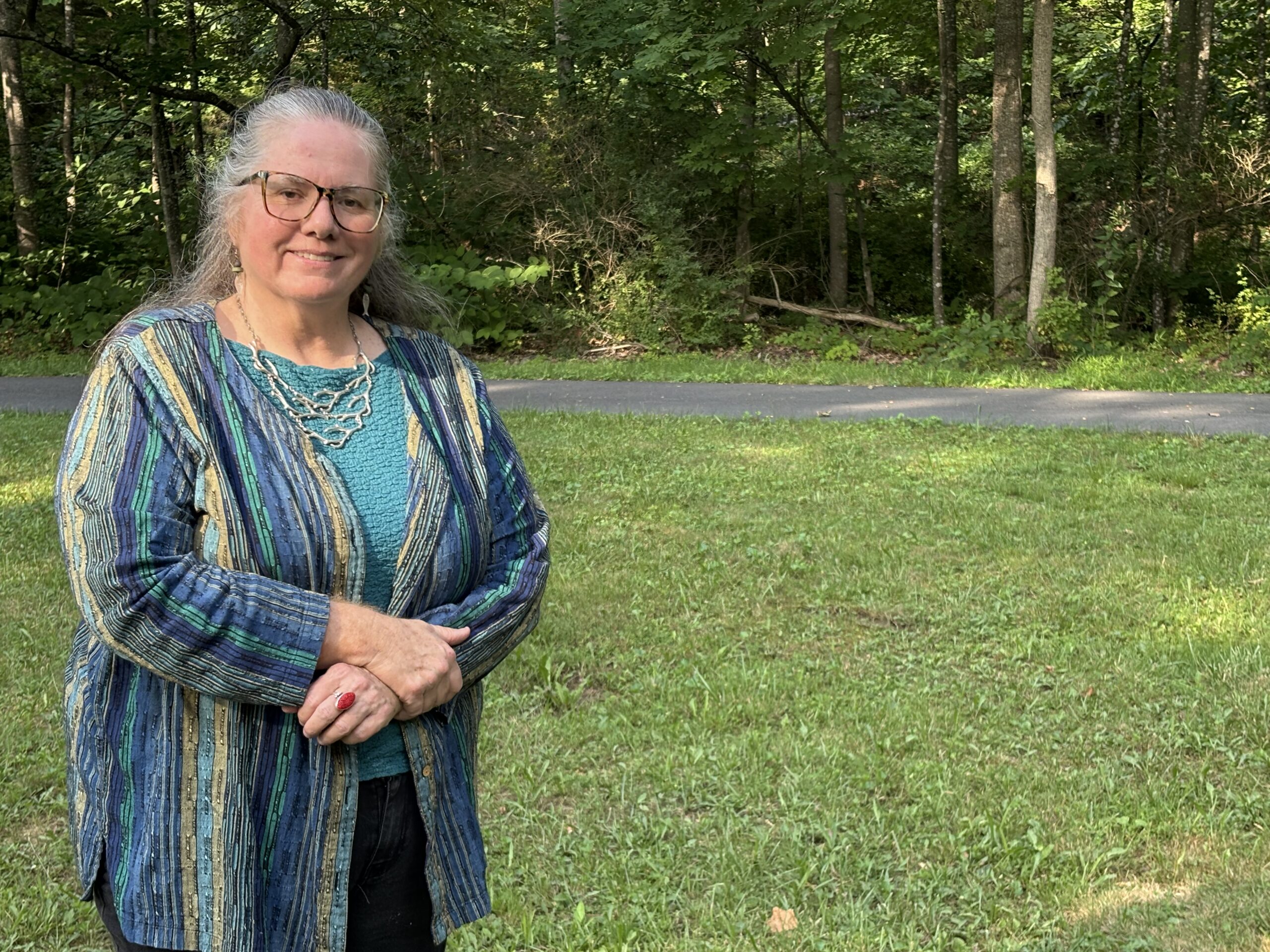 Woman in a colorful striped cardigan stands outdoors, smiling, with a natural backdrop of trees and grass. The image captures a serene moment in a park.